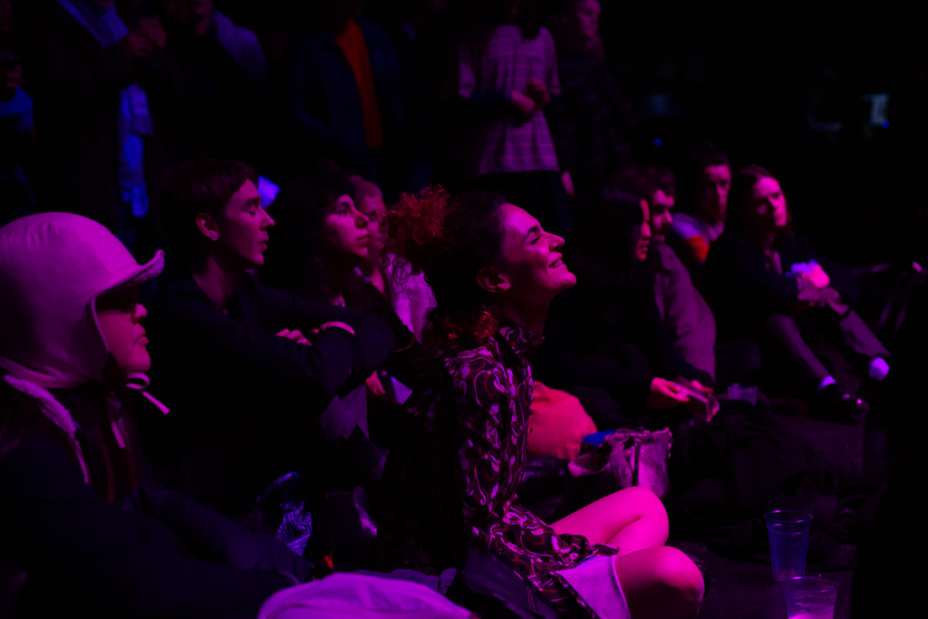 An audience sit and stand in a dark room with a wash of pink light highlighting faces. The image focuses a person sitting with head tilted upwards, smiling, eyes closed, listening intently