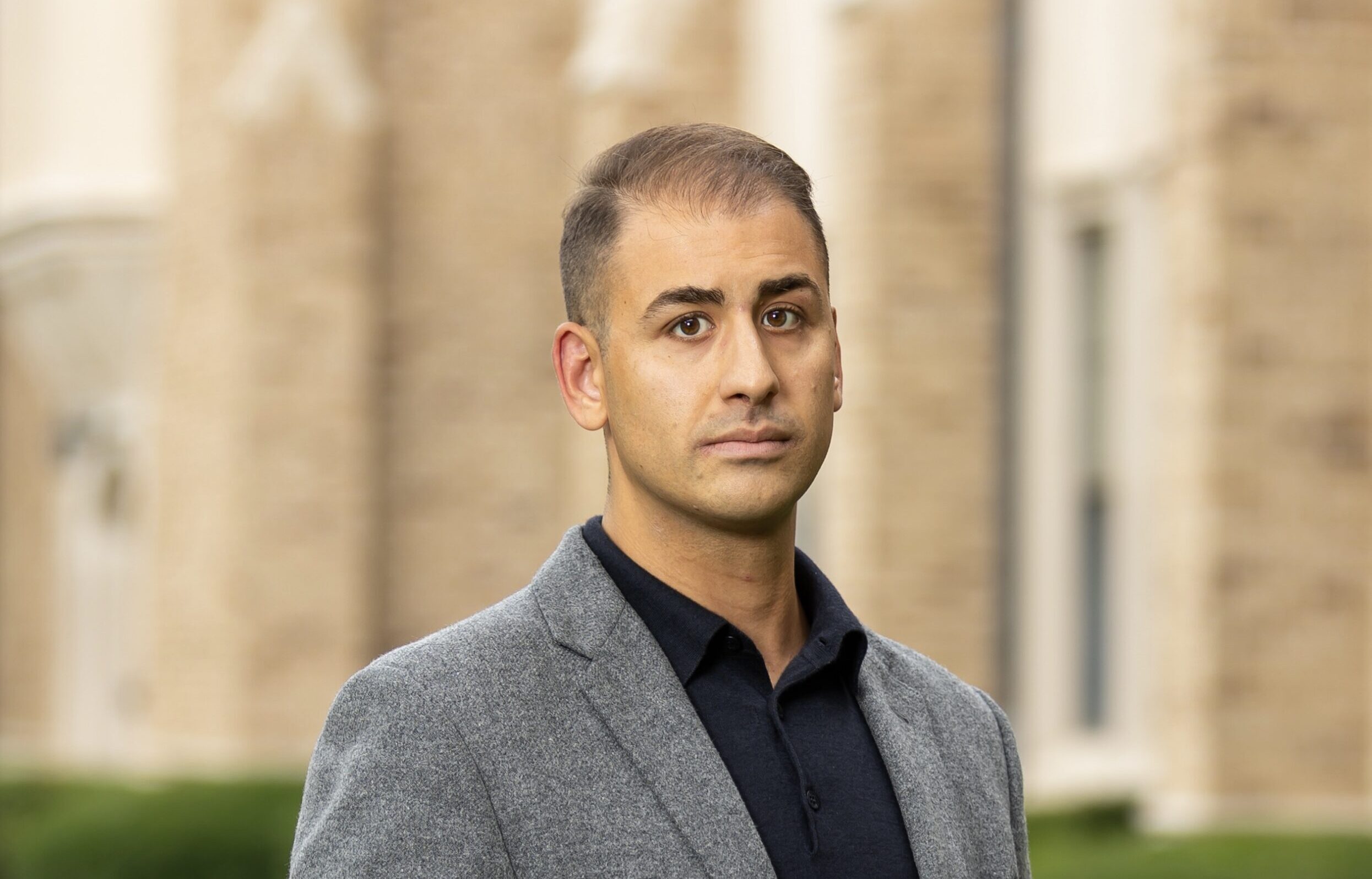 A photo of Peter, a man with light skin brown hair and dark eyes. He wears a black shirt and a grey blazer