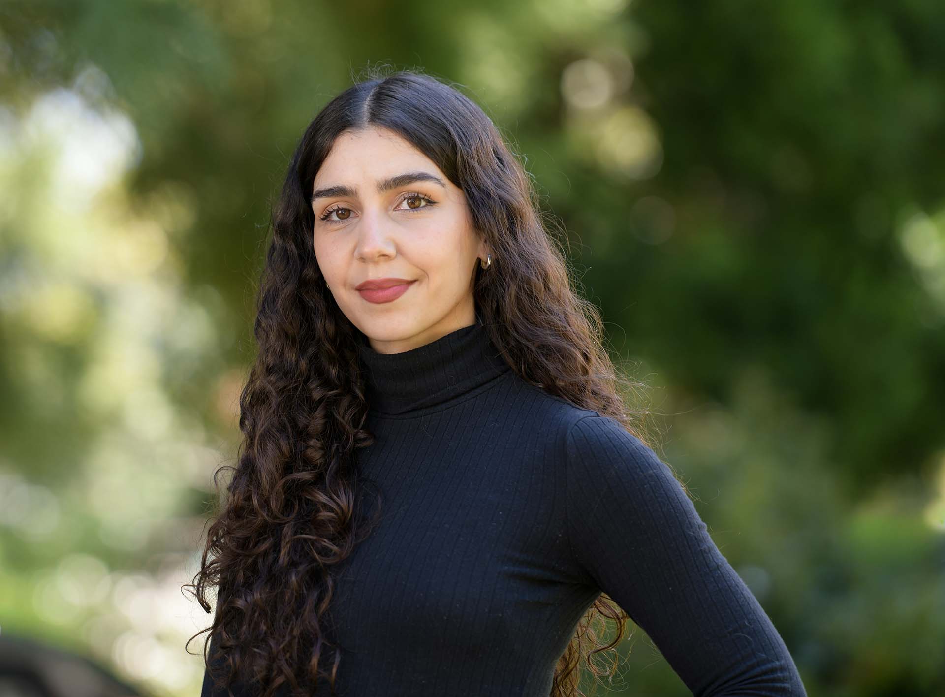 A photo of a West Asian woman with olive skin, curly dark brown/black hair and brown eyes. She is looking directly at the camera, posed with one arm resting on her hip. Her hair is centre parted and falls to her waist and she wears a navy blue turtle neck, soft red lipstick, black mascara and silver earring. The photo is well lit by natural daylight and the background of the image is unfocused foliage and trees.