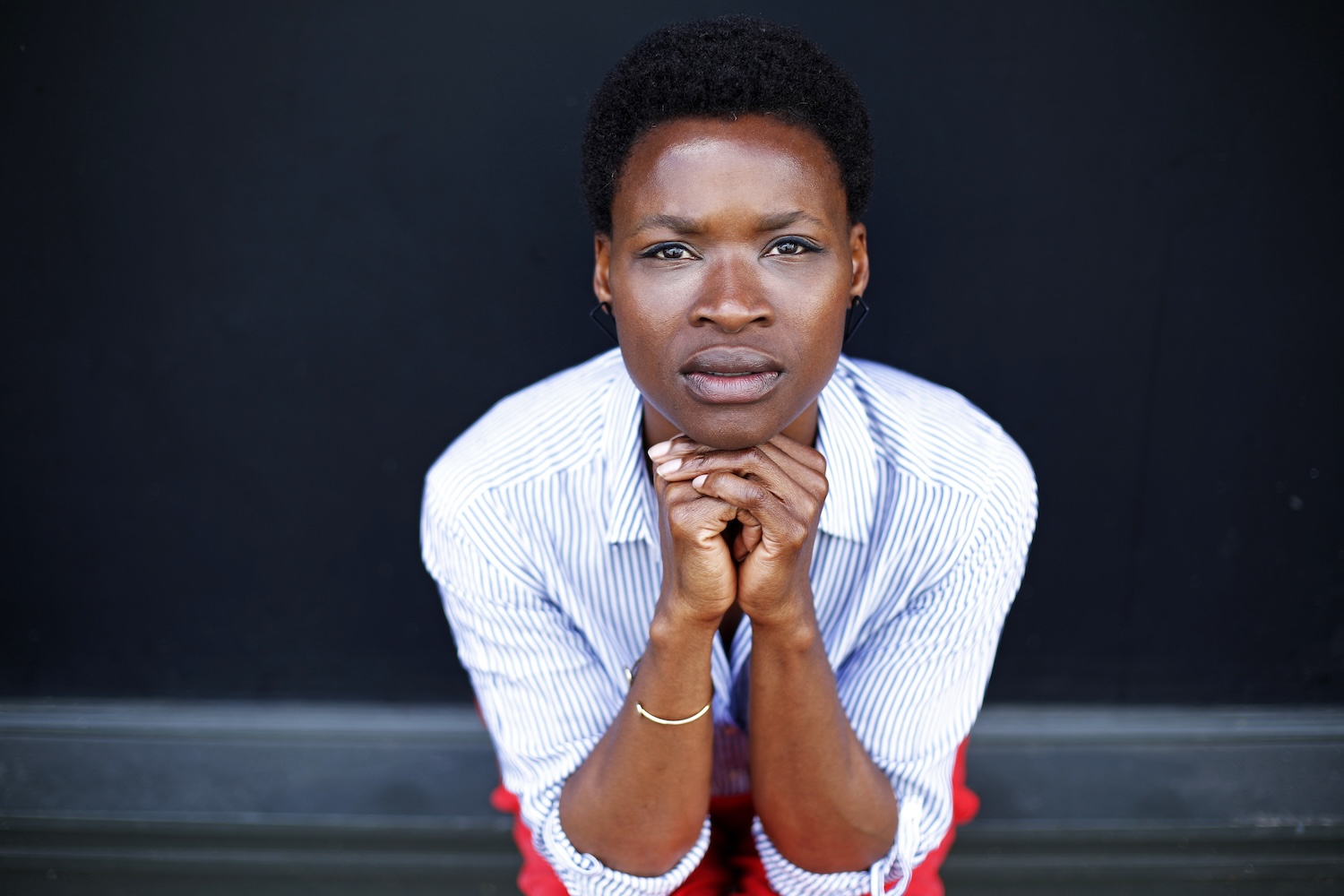 A headshot of Adura Onashile posed close to the camera with her hands clasped under her chin, making eye contact with the camera. Adura is a black woman with dark brown skin and eyes, and dark brown hair in a short natural style. She wears a pin striped white and blue shirt, orange trousers, small black geometric earrings and a single gold band bracelet.