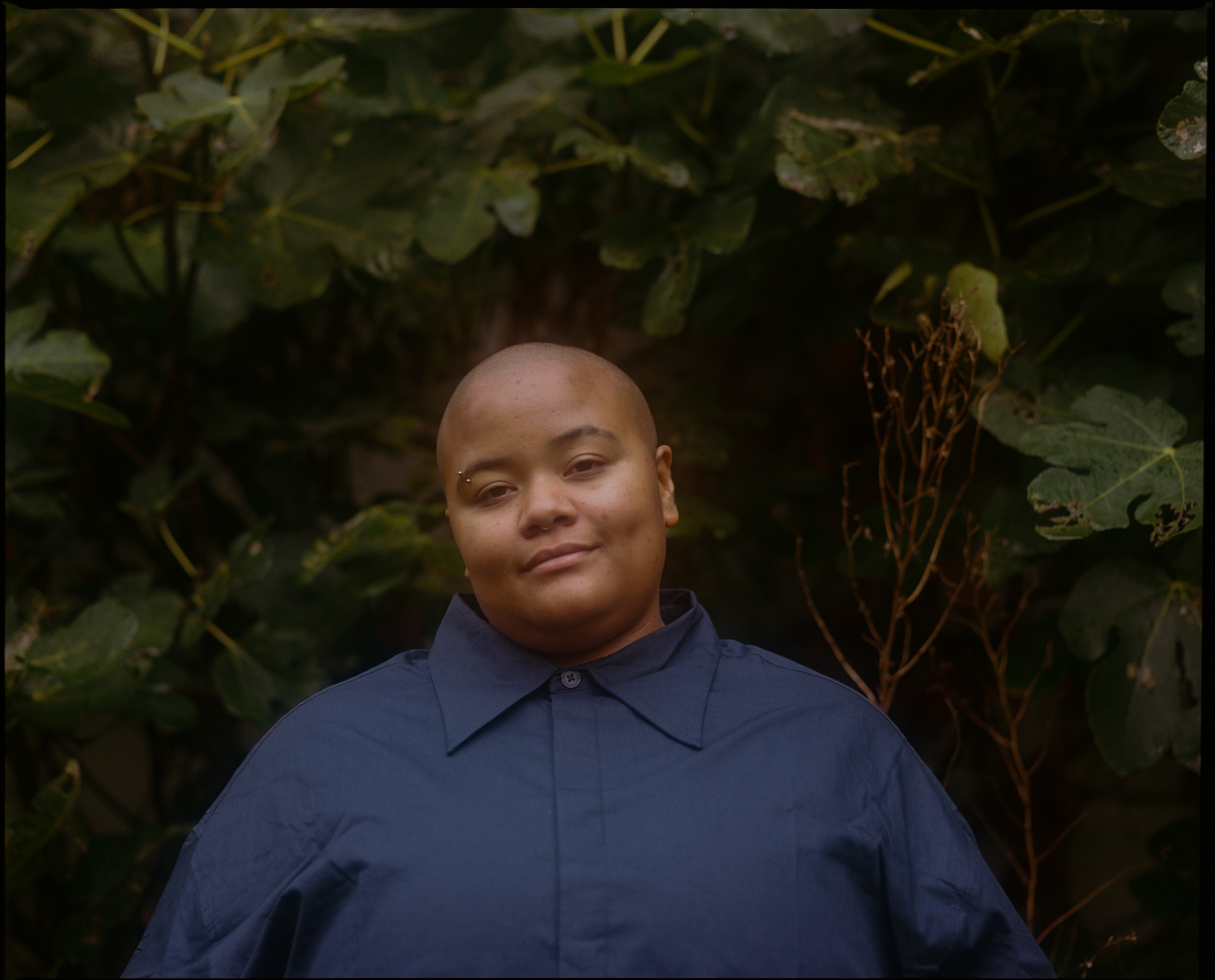 Nene is smiling against a background of green plants, they have a bald head and are wearing a dark blue collared shirt.