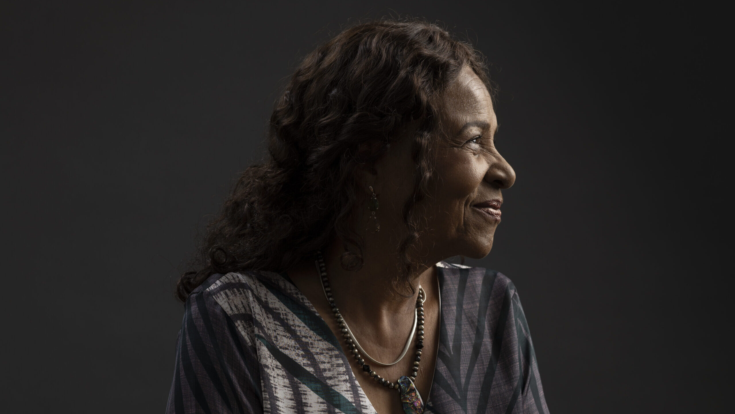 A headshot photo of Leda an older black woman with long curly hair. She wears a dark blue and beige top and simple necklace.